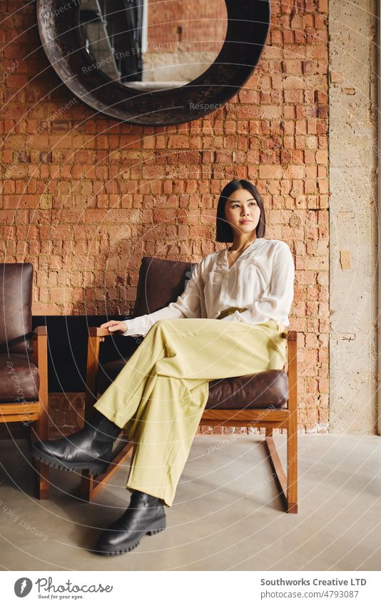 Portrait of confident mid adult Chinese businesswoman sitting on retro chair in office with exposed brick wall interior portrait chinese asian one people
