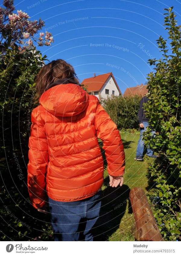 Boy with long hair and down jacket in bright orange with blue sky and sunshine in spring in a garden in Wettenberg Krofdorf-Gleiberg near Giessen in Hesse, Germany