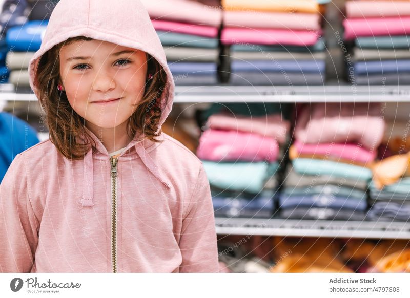 Little girl in clothes shop shelf smile hoodie casual customer glad portrait cute kid positive retail friendly child little apparel store garment dark hair pink