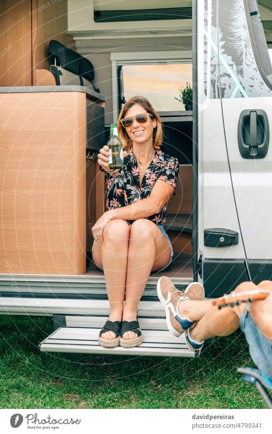 Woman showing beer bottle having fun with friends sitting at the door of their camper van smiling woman happy talking one drink sunglasses trip enjoying