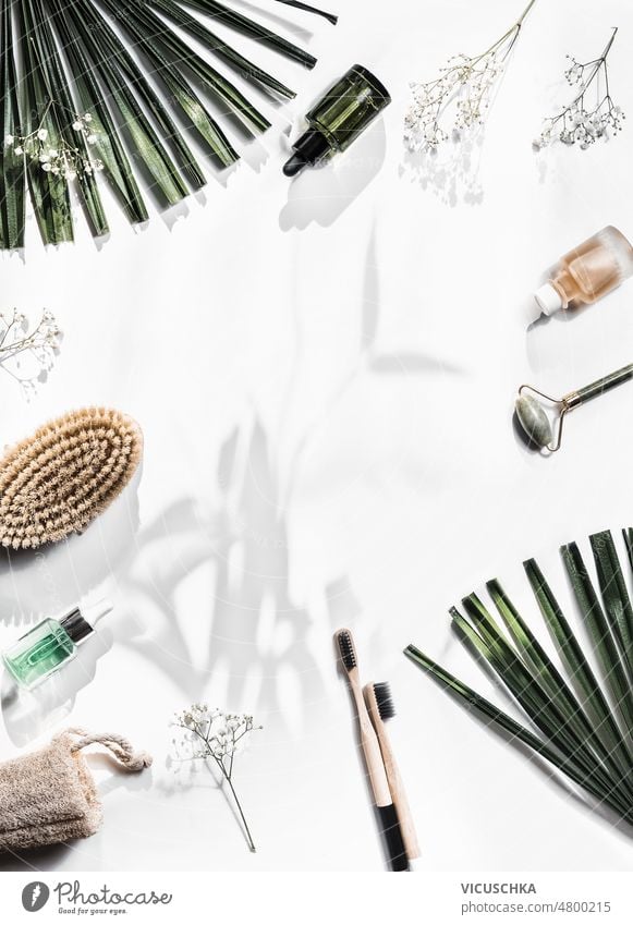 Sustainable beauty background with massage brushes, bamboo toothbrushes, cosmetic bottles, jade roller and sponge on white background with palm leaves