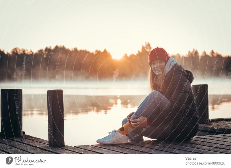 Young brunette with a beautiful, realistic smile sits on a wooden bridge by a lake at sunrise. Red hat with black coat and white shoes. Fashion style. Paltamo, Finland. Discovering scandinavia