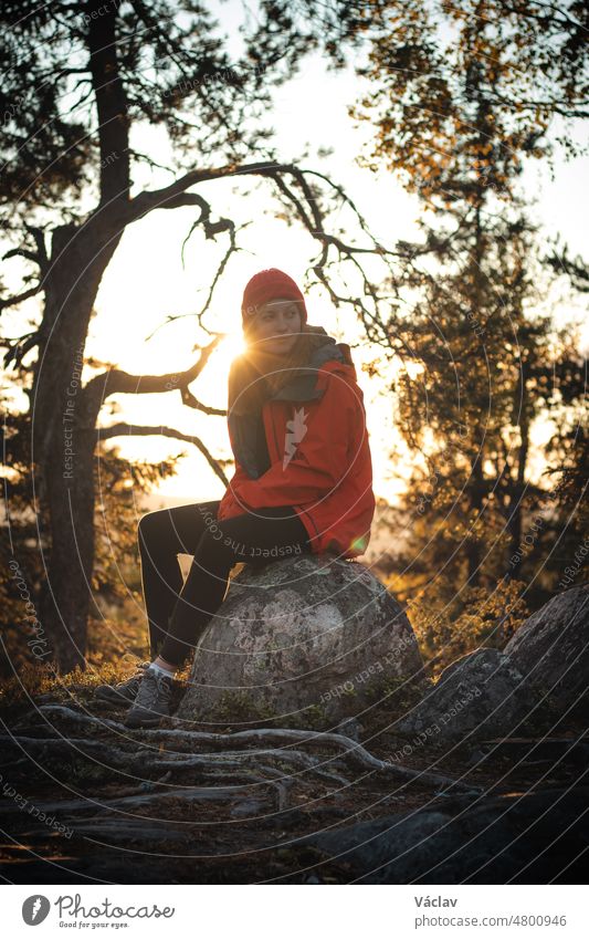 beautiful brunette traveller is sitting on top of a hill on a rock, resting after a hard climb. Sunset on top of a mountain in Vuokatti, Kainuu region, Finland. Natural smile of a girl in her 20s