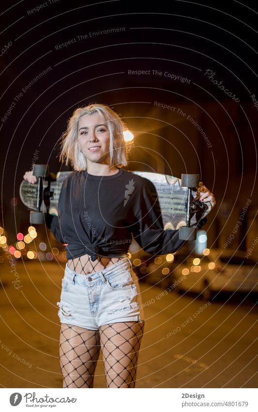 young pretty woman looking camera in shorts while standing on the street holding a skateboard at night in the city front view sensual long board girl