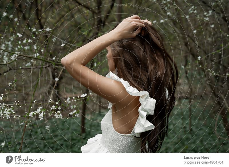 A brunette girl with long hair and a white dress is wandering in these woods. It’s kinda dark and moody image. And you can’t really see the girl's face. Blossom