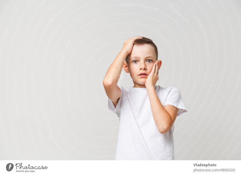 Terrified boy in white t-shirt with both hands on head on gray background horrified terrified scared kid child afraid fear person young emotion expression male