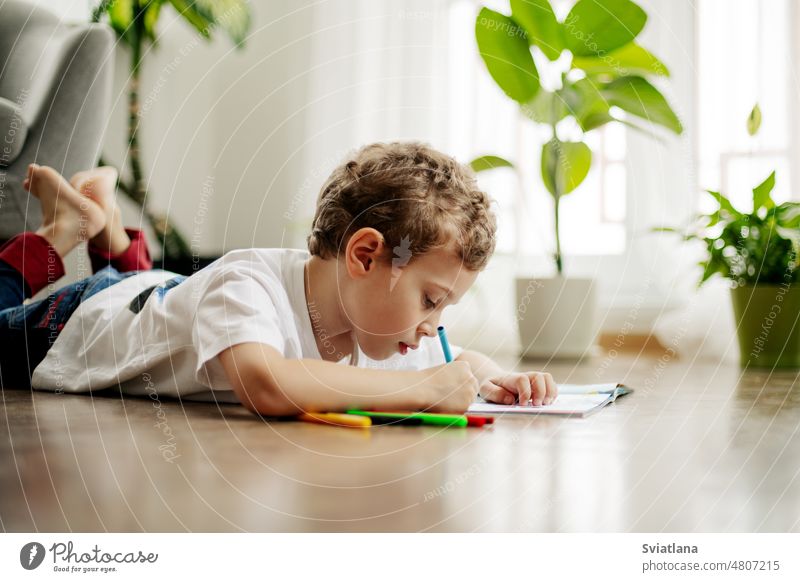 A little boy draws, lying on the floor, having fun. Happy childhood, positive emotions. Side view, space for text drawing home paint paper picture concept