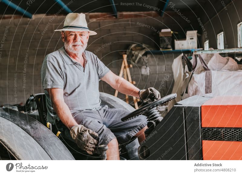 Aged male farmer sitting on tractor in garage man portrait senior countryside agriculture machinery aged rural vehicle shabby glove hat sunhat gray beard