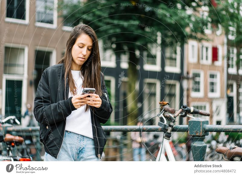 Focused young Hispanic woman using smartphone on bridge over canal message traveler vacation city trip mobile bicycle female hispanic ethnic amsterdam