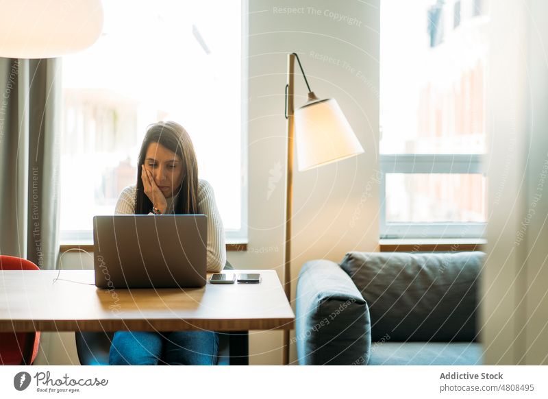 Focused young ethnic woman sitting at table with laptop and smartphones freelance thoughtful online hand on cheek work connection remote watch analyze focus
