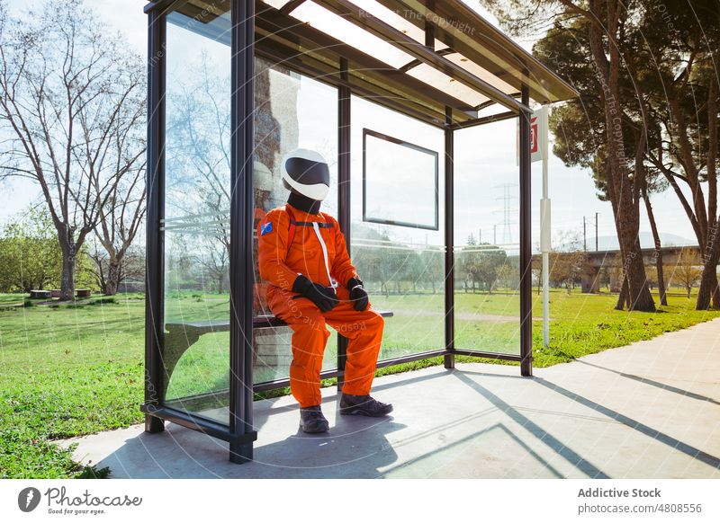 Unrecognizable cosmonaut in orange suit sitting at bus stop astronaut wait spacesuit public futuristic station bench spaceman mission expedition passenger male