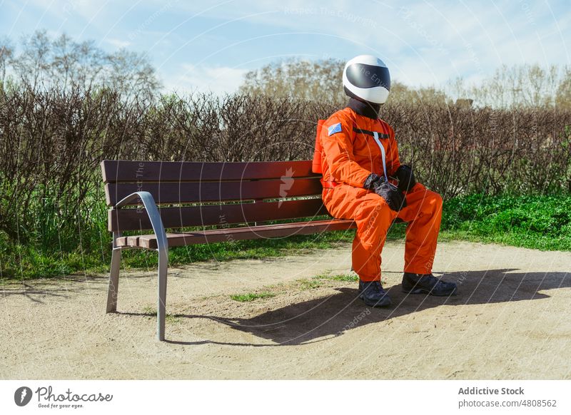 Unrecognizable spaceman in protective helmet and costume sitting on bench astronaut park cosmonaut nature futuristic spacesuit wait safety rest male