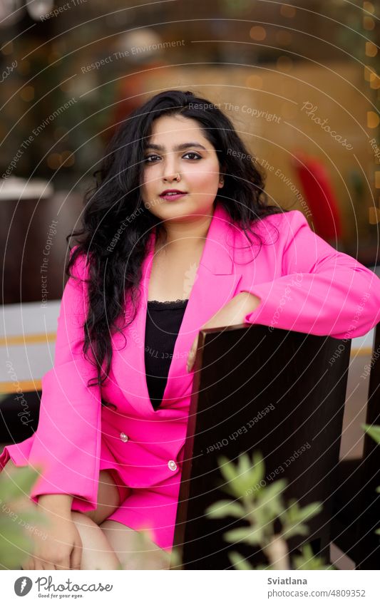 Portrait of a beautiful, attractive and elegant young Indian girl in a pink jacket sitting at a table in a cafe smiling indian portrait style glamour