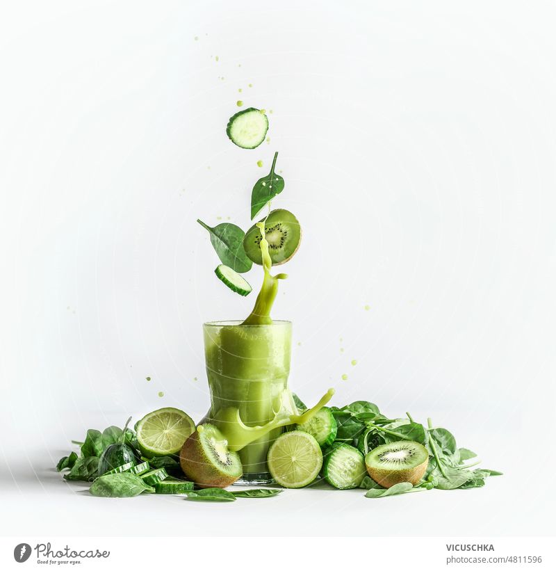 Splashing of green smoothie in glass with and flying ingredients at white background with heap of green fruit and vegetable cucumber detox drink fresh freshness
