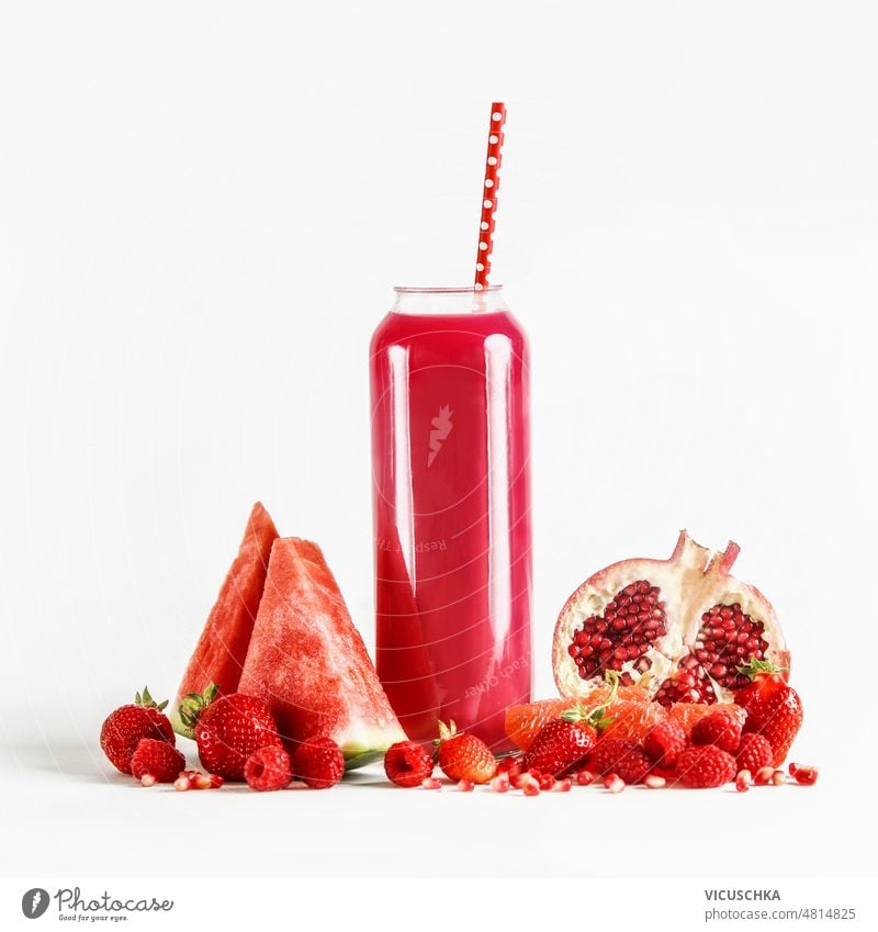 Red smoothie in glass with drinking straw at white background with fruit ingredients watermelon pomegranate strawberries raspberries healthy refreshing drink