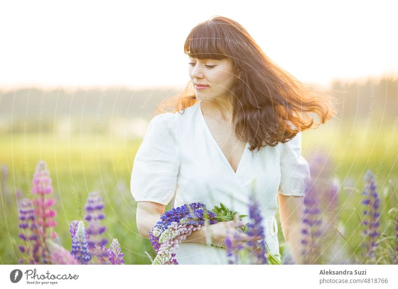 Beautiful woman in white sundress enjoying the summer nature. Picking colourful flowers, breathing fresh air and floral scent, walking in sunny field of lupins, wind in the hair. Happiness concept