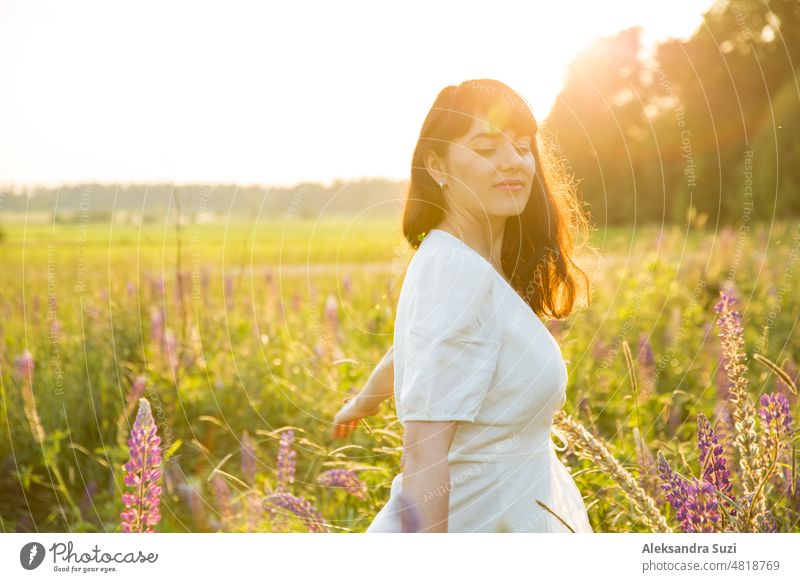 Beautiful woman in white sundress enjoying the summer nature. Picking colourful flowers, breathing fresh air and floral scent, walking in the sunny field of lupins. Happiness concept