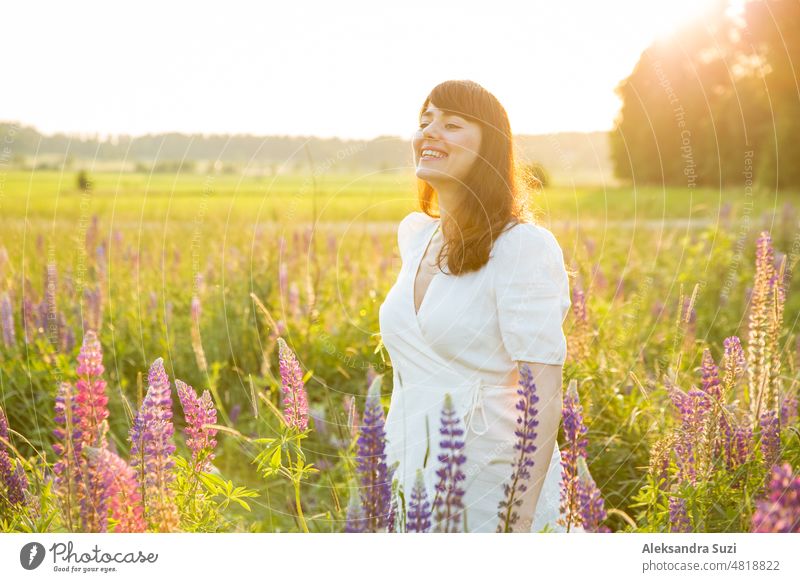 Beautiful woman in white sundress enjoying the summer nature. Picking colourful flowers, breathing fresh air and floral scent, walking in the sunny field of lupins. Happiness concept
