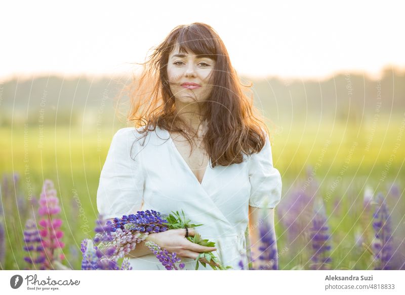 Beautiful woman in white sundress enjoying the summer nature. Picking colourful flowers, breathing fresh air and floral scent, walking in sunny field of lupins, wind in the hair. Happiness concept