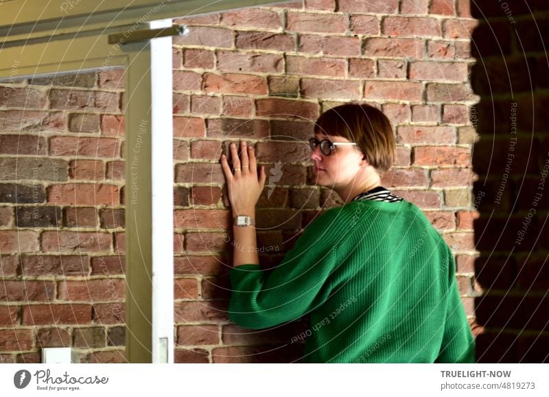 [Hansa BER 2022] Always along the wall seems to go: a young, retro styled woman with auburn bangs hairstyle, glasses, look to the left, wristwatch, green XXL cardigan in also color harmonious brick wall contact