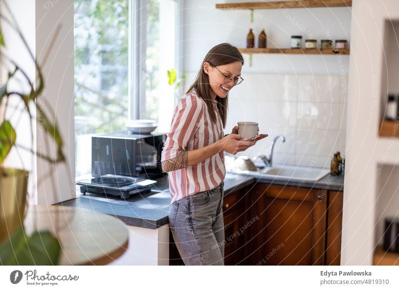 Young woman using smartphone while standing in her kitchen at home mobile phone technology online internet using phone communication domestic life confidence