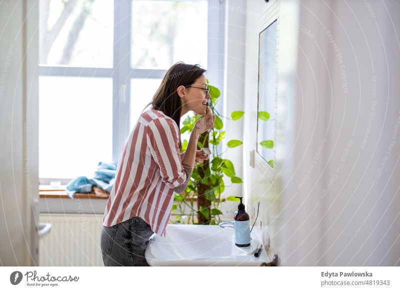 Woman applying lip gloss in front of the bathroom mirror domestic life confidence woman indoors home house people young adult casual female Caucasian attractive