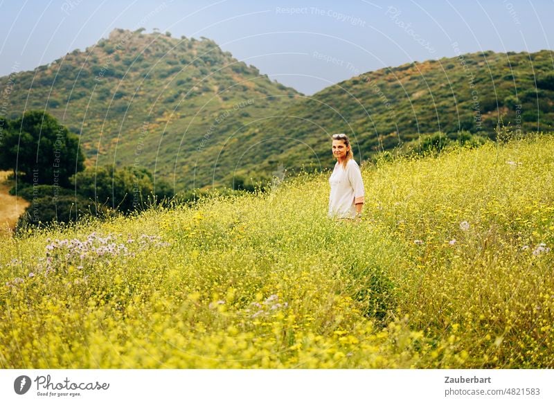 Woman in white sweater stands on dirt road in yellow sea of flowers in front of hill range off the beaten track Yellow Broom Hill chain of hills look Hiking