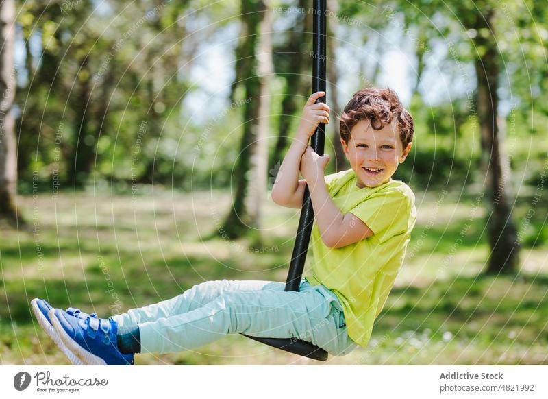 Cheerful little kid on swing rope on playground smiling at camera child smile childhood cheerful activity boy park positive portrait hang excited joy summer