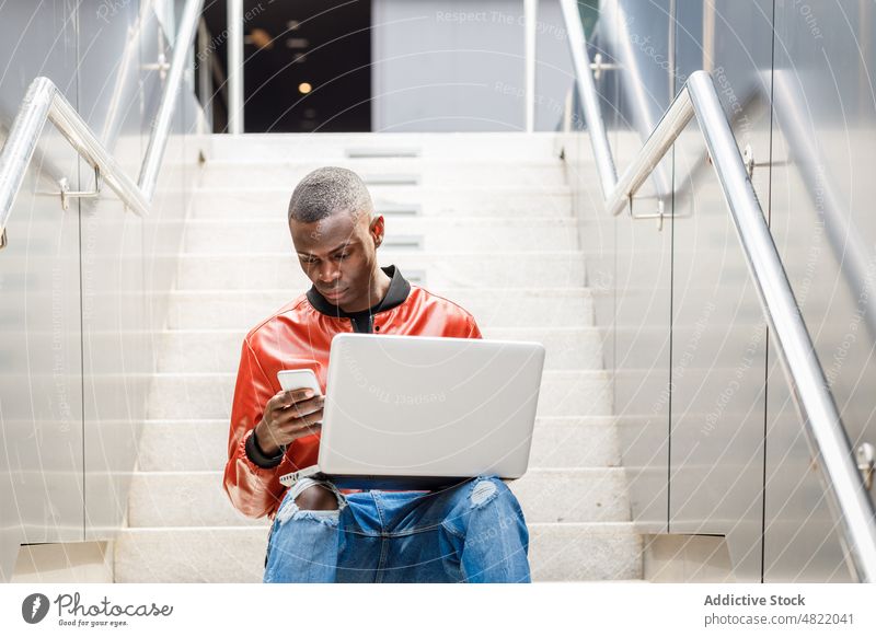 Self employed black man on smartphone and working on laptop on street stairs freelance remote city multitask male young african american ethnic trendy