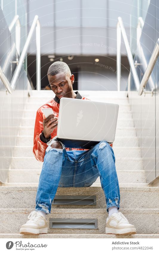 Self employed black man on smartphone and working on laptop on street stairs freelance remote city multitask male young african american ethnic trendy