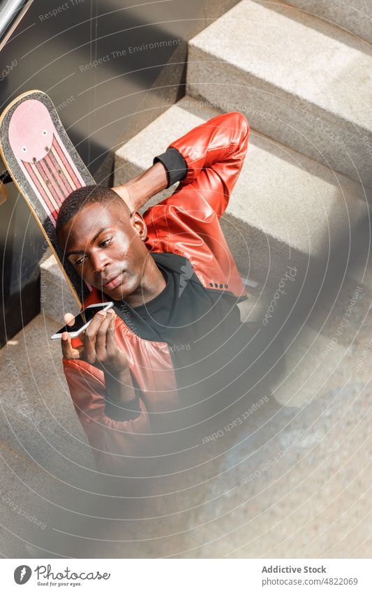 Trendy black guy sending voice message on smartphone resting on stairway leaning on skateboard man relax audio phone call street city skater talk