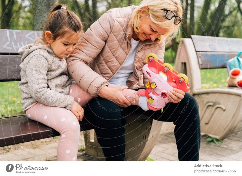 Smiling grandmother putting roller skates on feet of little girl in park woman child put on positive help granddaughter together activity hobby bench casual kid