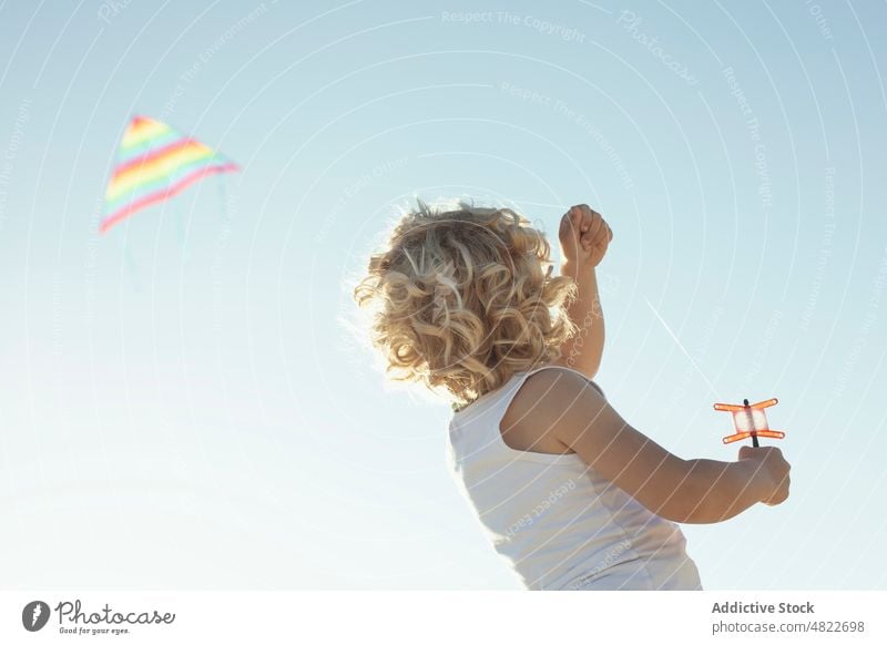 Anonymous child launching colorful kite summer childhood vacation boy blue sky cloudless joy kid blond curly hair sunlight positive activity hobby holiday
