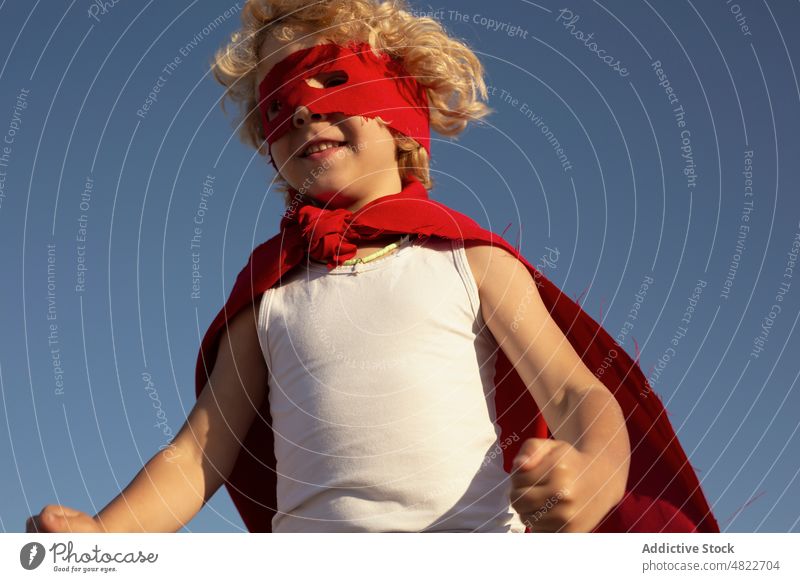 Happy little superhero smiling under blue sky child happy smile character brave clench fist positive boy costume excited cape mask pretend kid blond curly hair