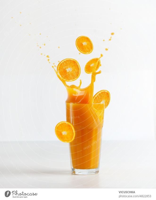 Glass with splashing of orange juice and falling orange slices  at white background glass table healthy refreshing drink liquid motion front view beverage