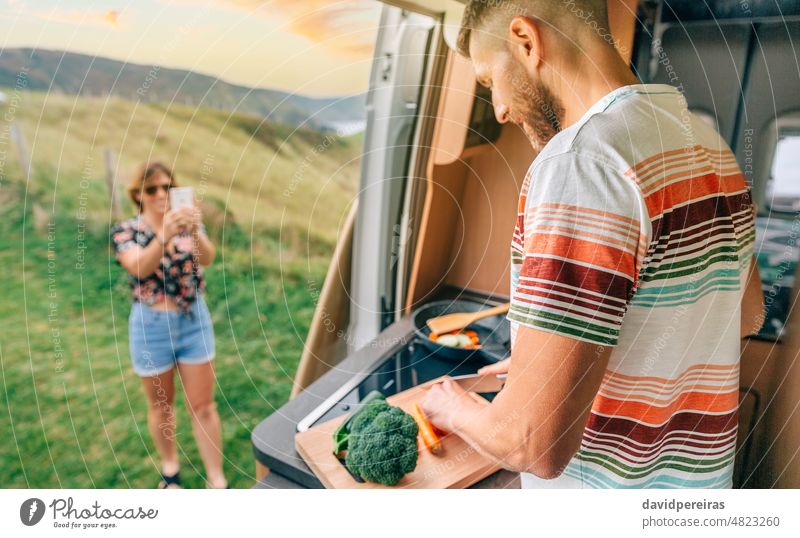 Young man cooking vegetables in a camper van while his wife takes a picture of him with her cell phone young mobile woman vegan friend couple vegetarian cutting