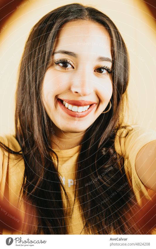 Young brunette girl over isolated yellow background taking a selfie and showing tongue to the camera while having fun. Happy woman. People sincere emotions lifestyle concept