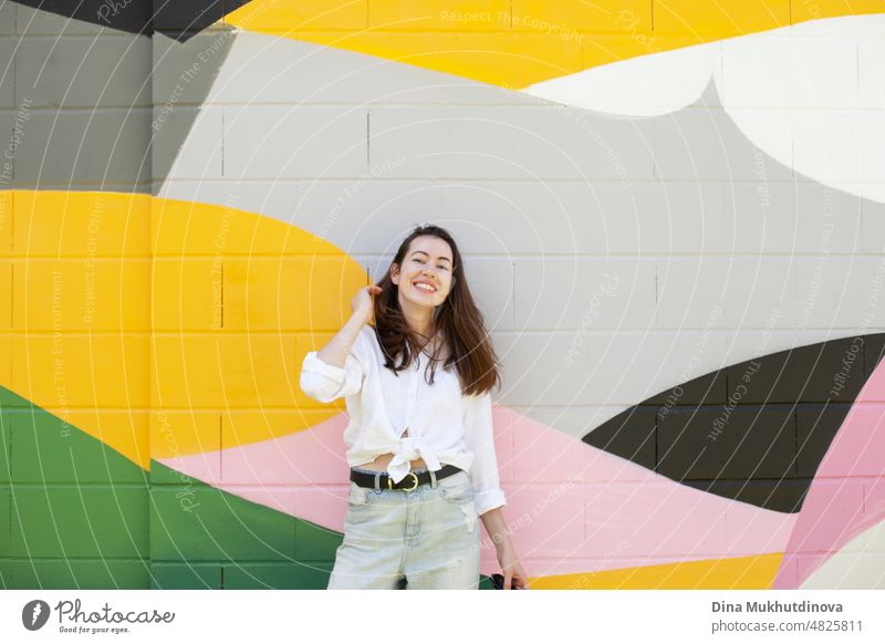 Young woman in white shirt and light blue jeans standing near colorful vibrant wall. Candid lifestyle summer style portrait. beautiful city happy urban girl