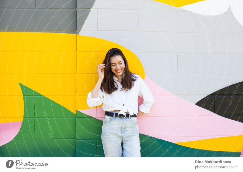 Young woman in white shirt and light blue jeans standing near colorful vibrant wall. Candid lifestyle summer style portrait. beautiful city happy urban girl