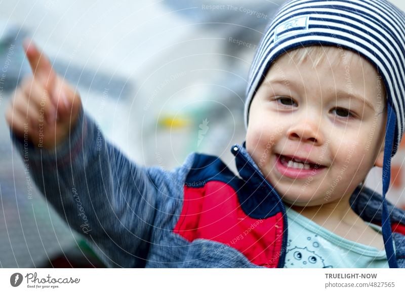 Hehe - that's the way! Little man of one and a half years with striped cap, blue-red jacket and T-shirt already knows best, smiles, beams, shows first little teeth and stretches out arm and index finger