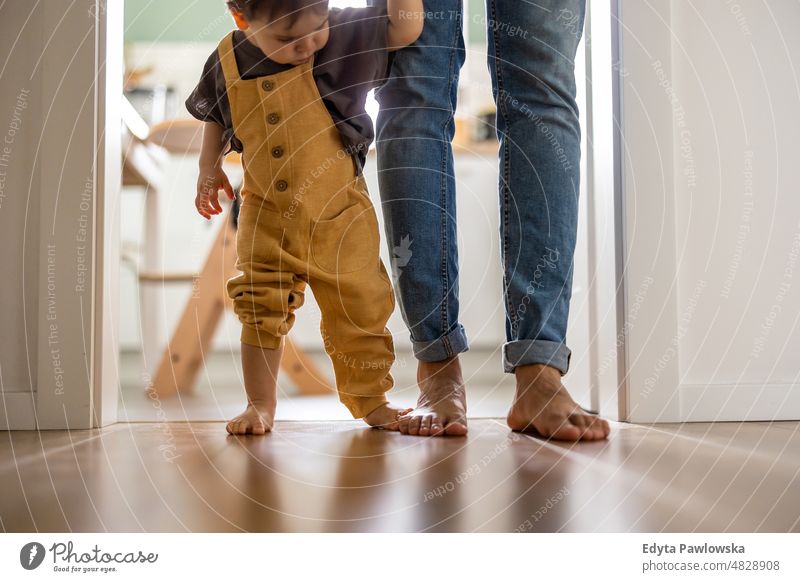 Little baby boy learning to walk with his father beside him at home walking standing active adult affection barefoot bonding candid care casual cheerful child