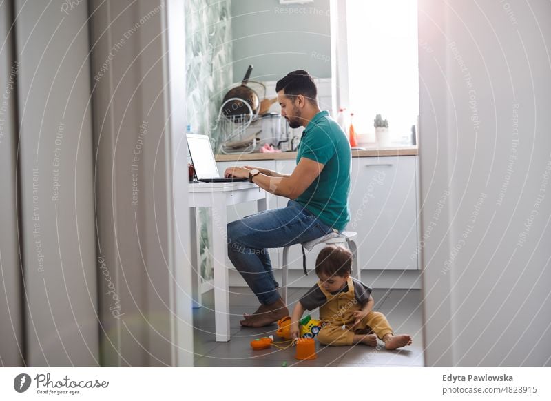 Father working on laptop while child playing on kitchen floor adult affection baby bonding boy candid care childhood domestic enjoying family fun happiness