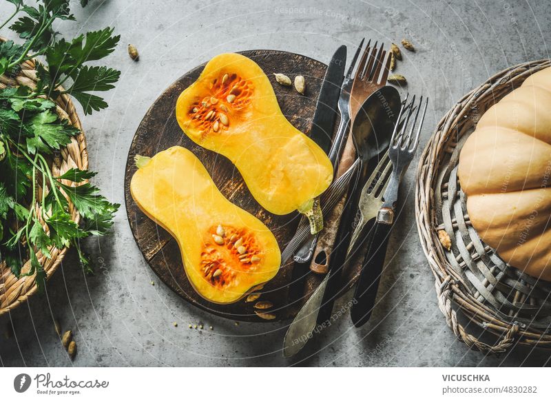 Halved butternut squash on plate with cutlery and herbs at grey concrete kitchen table background. halved cooking home seasonal vegetables top view autumn food
