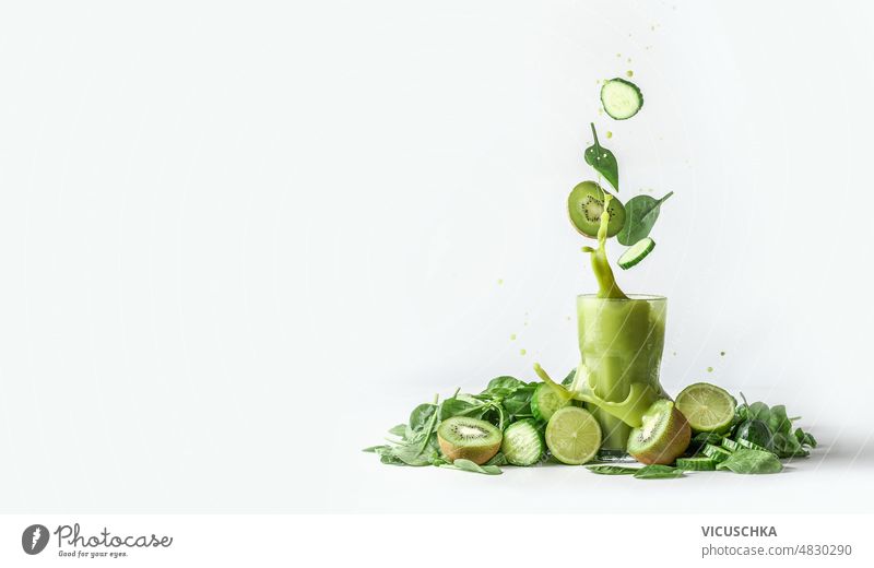 Green smoothie splashing in glass and flying ingredients at white background with heap of green fruit and vegetable cucumber kiwi spinach leaves border
