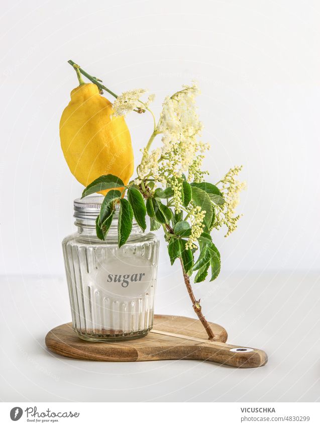 Elder flower syrup ingredients: Elder branch, lemon and sugar in glass jar elder flower cutting board white background front view blossom brown
