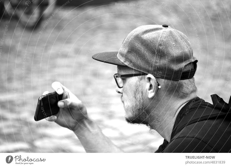 [Hansa BER 2022] Portrait Young man with glasses and baseball cap looks intently at his smartphone, which he holds in his right hand in front of him to take pictures. b/w black and white