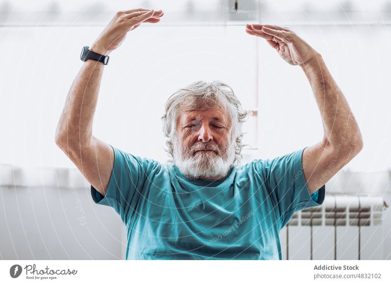 Senior male meditating with raised arms man meditate yoga home morning zen window spirit session balance wellbeing practice relax mindfulness eyes closed
