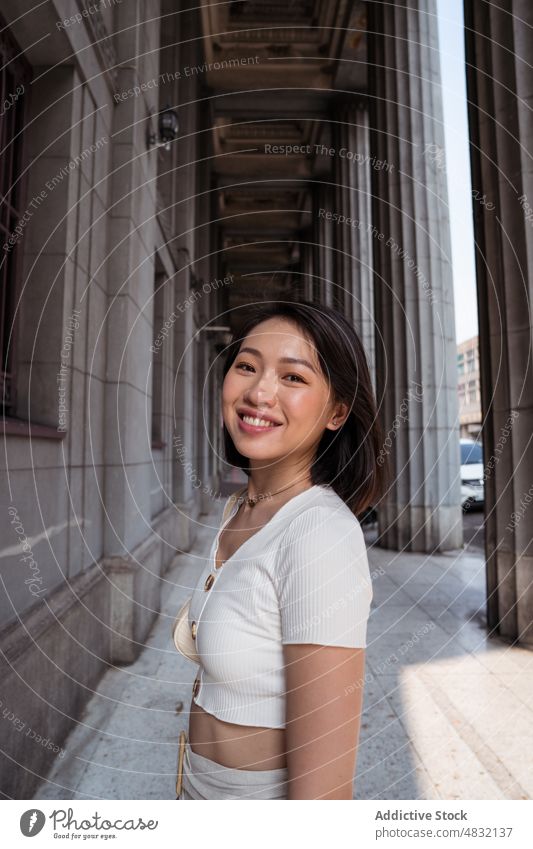 Content young Asian lady standing near building with columns in sunlight woman smile passage vacation sightseeing happy tourist positive style confident female