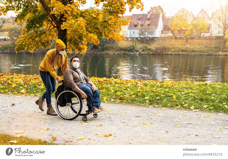 Woman and her friend with face mask in a wheelchair having stroll through the park at a river enjoying the autumn during covid-19 pandemic disability city