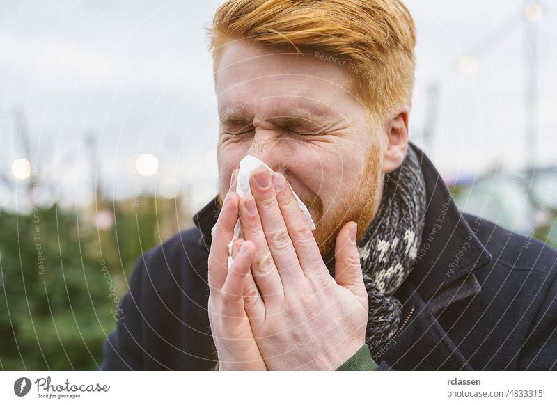man sneezes and blows his nose in a handkerchief at winter time with allergy or an infection a virus. Portrait of redhead man outdoor. sinus adult allergic